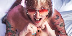 Kate Bornstein laying on a bed using her arms to prop up her upper body to pose for the camera with vibrant facial expression. Kate is a white person with short blonde hair. She is wearing tinted colourful glasses, has brightly coloured fingernails and has colourful tattoos on her legs and arms.