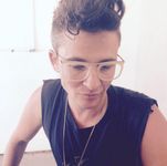 Sam Feder looking down in a calm and candid photograph. They are dressed in a sleeveless black shirt with a frayed edge, and a long gold necklace around their neck. The background of the photograph is overtaken by a strong wash of overexposed light.