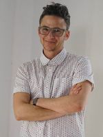 Sam Feder smiles while standing with arms crossed in a casual but confident pose in front of a white wall. Sam is a white person with short dark hair and glasses. They are wearing a short-sleeved, light-colored button-up shirt with a subtle pattern and a wrist watch.