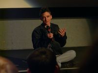 Sam Feder sitting cross-legged on a stage floor and talking to the audience through a microphone during a post-film discussion period. Sam is a white person with short dark hair and glasses. They are wearing a dark jacket and light-colored pants.