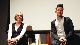 Kate Bornstein and Sam Feder are equipped with microphones and seated on stage after a film screening as they engage with the audience in discussion. Kate, an older white person with an ear-length bob, smiles and listens attentively. She is wearing a long white sleeve top with a black vest on top. Sam, a white person with dark coiffed hair, also engages with the speaker that is off screen. They are wearing a subtle patterned button up shirt with a dark cardigan on top.