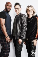 Full body studio portrait of Marquise, Sam and Amy in a group standing pose. Marquise Vilson, a Black person with dark facial hair and bald head, stands on the left. He has a muscular build and is wearing a black shirt with a dark vest and dark patterned pants. Sam Feder, a white person with dark coiffed hair and glasses, stands in the middle and wears a leather jacket and dark pants. Amy Scholder, a white person with shoulder-length light hair and glasses, stands on the right. She is wearing a dark sweatshirt and dark pants with a gold necklace.