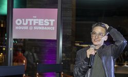 Candid photo of Sam Feder speaking into a microphone at the happy hour bar venue. Sam is a white person with dark coiffed hair and bold glasses. They are wearing a leather jacket on top of dark clothing.