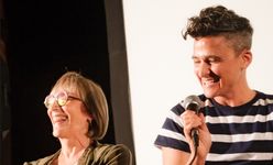 Candid photograph of Sam Feder and Kate Bornstein sharing a laugh together on stage. Sam Feder, a white person with dark coiffed hair, is wearing a striped shirt and holds a microphone. Kate Bornstein, a white person with a short bob and round glasses, laughs beside them.