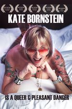 Kate Bornstein lies on her stomach on a bed with her chin propped on her knees and legs in the air. Kate is a white person, has a blond bob, and wears aviator sunglasses. Kate wears socks and is topless, showing off her tattoos and punk jewelry.