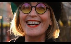 Close up joyful portrait of Kate Bornstein, an older white person with light skin and shoulder-length blonde hair. She is wearing a dark hat along with her her iconic colourful round glasses and silver nose and lower lip piercings. The background is soft and blurred red and brown tones. A beam of golden-hour sunlight illuminates Kate's hair.