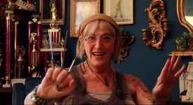 Still of Kate Bornstein in a decorated living room, excitedly talking to the camera while holding three butterknives between her fingers, which are curled in a fist. Kate is an older white person with light skin and shoulder-length blonde hair. The room has dark walls and golden accents. Behind Kate is a shelf holding several awards and tiaras.