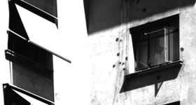 A black and white filter over an image of a pale stone building. A few windows are visible. The left ones have small awnings over them, and the one window on the right is open with curtains blocking the inside of the room.