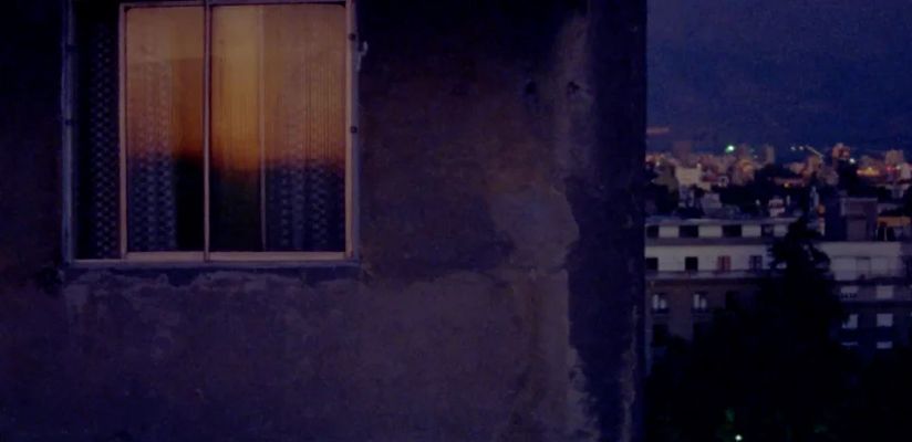 The edge of a building occupies the left two-thirds of the image. There is a visible window with light coming through but the shades are blocking any view into the window. The right two-thirds of the image features a skyline at night. The image has a blue tint over it due to the time of day.