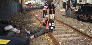 A person with light tan skin sits on the metal edge of a train track in an urban environment. They are wearing a tan baseball cap backwards, a black jersey jacket with a pink t-shirt underneath, black denim pants, and light pink and light blue shoes. There is a person slightly in front of them, hunched over taking a picture of the sitting person with a black camera that is in their hands. The camera-person has dark brown skin, is bald, and is wearing a black puffy sleeveless vest over a white and light-gray-striped long-sleeve t-shirt. In the background, three other people stand around watching the sitting person.