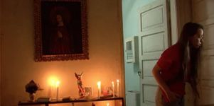 A person peers through a slightly open door. They have long straight brown hair, light skins, glasses, and are wearing a red short-sleeve t-shirt and light denim jeans. The room the person is standing in is dimly lit in orange and yellow by six candles sitting on a stand pressed against the wall. To the person's side is an open door into a room which is much more brightly lit, in a greenish hue. Above the candle-stand is a painting of a person with their hands together praying. The person in the painting has light skin and wears a red garment. The painting is held in an ornate gold frame.