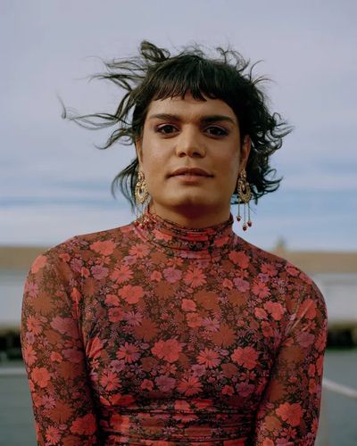 Zaiba Baig has shoulder length hair escaping in the wind, brown skin, a slight smile and a direct gaze. They wear a coral top with a floral print and large gold earings. The background is blurred but suggests a dock or pier.