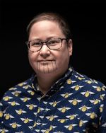 Theo Jean Cuthand wears glasses, has short hair, and is a light-skinned person of Plains Cree and Scots descent. He has facial tatoos on his chin of dots and vertical lines. He wears a smart button-up with a print of bees.