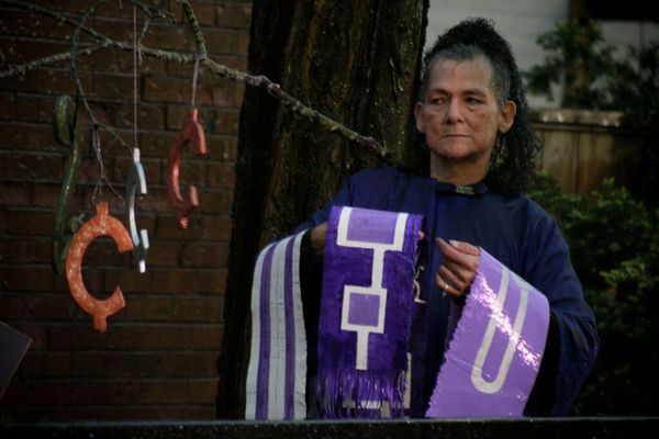 Still of trans Two Spirit Haudenosaunee performance artist Aiyyana Maracle in her 60s wearing a long deep purple robe and carrying three purple banners outside.