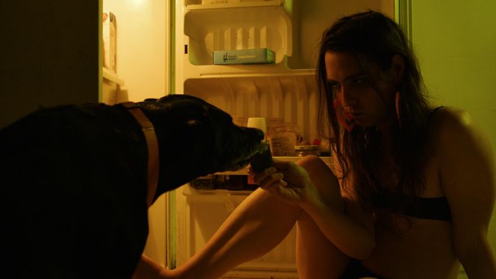 Illuminated by the light inside an open fridge door, a fair skinned trans woman sits on the floor and hand feeds a dog a piece of fruit. She is wearing nothing but undergarments and large pink heart earrings. Her brown hair reachers her chest and she appears slightly disheveled or intoxicated.