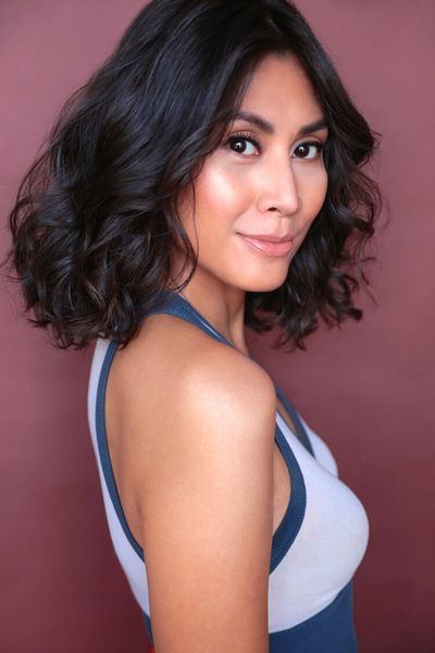Rain Valdez glances over her shoulder into the camera with a slight smile. Rain is an East Asian person with dark curly shoulder-length hair and light skin. She wears a white and blue tanktop and subtle makeup.