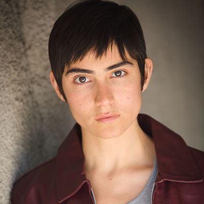 Izzi Rojas with a neutral expression. Izzi is a Latine person with light skin and short dark hair. They wear a dark red leather jacket.