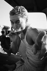 Black and white photo of Lío Mehiel in a car, smoking, and pointing at the camera. Lío is a Puerto Rican and Greek person with straight chin-length hair. They wear a muscle top and jeans.