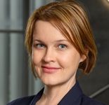 Phoebe Hart is white and intersex. She has light skin wearing a navy blue blazer shown from the shoulders up. She has a short blonde bob haircut and light blue eyes. The background is a blurred gray color.