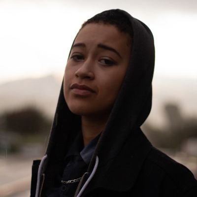 Savannah Ward is a Black trans/non-binary person with light brown skin in front of a blurred out landscape. They are wearing a black zip up hoodie pulled over their head. They are wearing a dark blue button up with a gold chain under the collar of the button up.