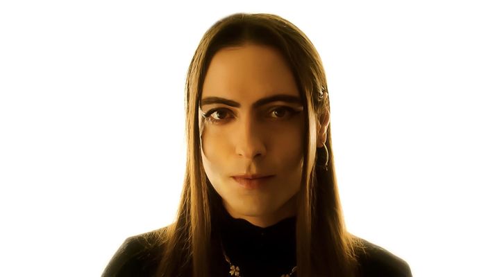 Sepand Mashiahof is a trans-femme Iranian-American in front of a bright white background. She has light skin and long straight brown hair. She is wearing a black turtleneck, a gold necklace and gold dangle earrings.