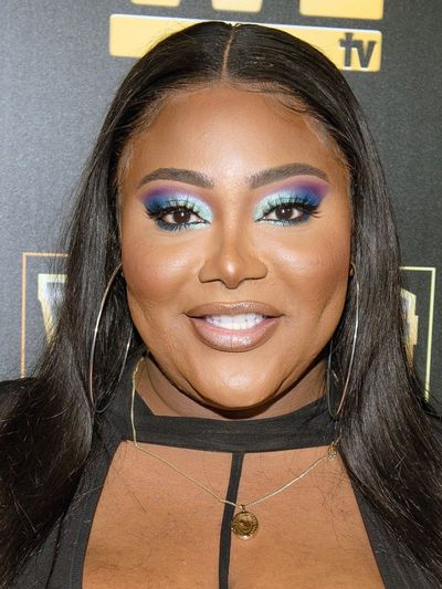 Ts Madison is a Black trans woman with brown skin in front of a black background. She has blue and purple eyeshadow with nude lipstick and long straight black hair. She is wearing big gold hoop earrings and a gold necklace. She's wearing a black top with the chest exposed.