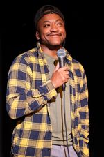 Kai Choyce is a Black trans man with brown skin and black hair. He is seen mid-standup performance, smiling into a microphone in front of a black background. He is wearing jeans, a backwards black baseball cap, and a yellow-and-black plaid sweater over an olive shirt.