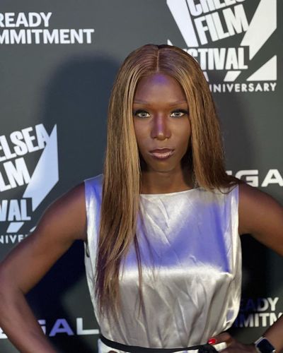 Daniella Carter is a Black trans woman with dark brown skin. She is posed in front of a film festival poster with long straight brown hair. She is in a silver metallic sleeveless dress.