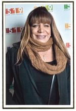 Monica Forrester is a Black trans woman with light brown skin, posed in front of a white banner. She is wearing a light brown infinity scarf and has long straight brown hair with bangs. She is wearing a black zip up jacket and a black t-shirt underneath.