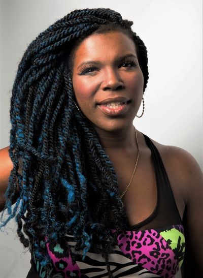 Mizz June is a Black trans woman with dark brown skin posed in front of a white background.  She has long black twists with blue accents and is wearing hoop earrings. She is wearing an assorted patterned shirt including a cheetah, zebra, leopard print in assorted colors.