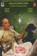 A medium skin toned South Asian adult man sits leisurely in a lounge chair and pets a large black dog with a silver chain on its neck. A chest of treasures is inserted at the bottom left corner near the man's other hand. A closeup of a ring on a finger is imposed on the bottom right corner of the poster. The poster has a green background with a starry sky illusion and there is a drawing of a Hindu figure face above a shining star. Various text is featured on the poster about the film title, director, languages available and festival participation.