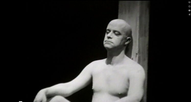 A fair skinned nude bald adult man sits on an empty black floor leaning back against a wooden beam. The person gazes down in a pensive manner. The still is in black and white.