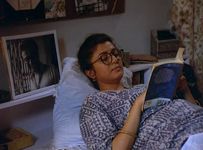 A South Asian young adult woman lays on a bed with her head propped up by a pillow to support her reading of a novel. The bedroom is decorated with a large framed sepia photograph of a person positioned near her pillow and various personal items.