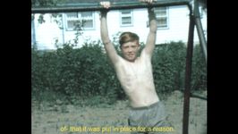 A young person with short orange hair is shirtless and in gray shorts holding onto a metal bar with their arms above their head. They are on dirt with bushes in the background. There is yellow text at the bottom that reads "of- that it was put in place for a reason."
