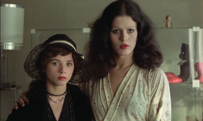 Two people stand next to each other. The person on the right is taller and resting their hand on the shoulder of the person on the left. The person on the right is in a white dress with red dots all over it with red lipstick, some black eyeshadow and thin eyebrows and shoulder length curly hair. The one on the left is wearing a black lace hat in a black outfit with a black beaded necklace and a pink and white beaded necklace.