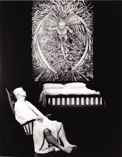Black and white image of a pale male figure in a dark bare room. The person is wearing nothing but a draped white sheet and is resting on a rocking chair with their eyes closed. Behind them there is a bed and a large angelic tapestry above the bed.