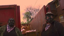Two people standing wearing gas masks in front of a house and a dumpster looking at the camera. The one on the left is wearing a camouflage flannel and a black t-shirt and a pink and blue patterned bandana around their neck. The person on the right has the same bandana and they are wearing a black zip up with a black t-shirt underneath.