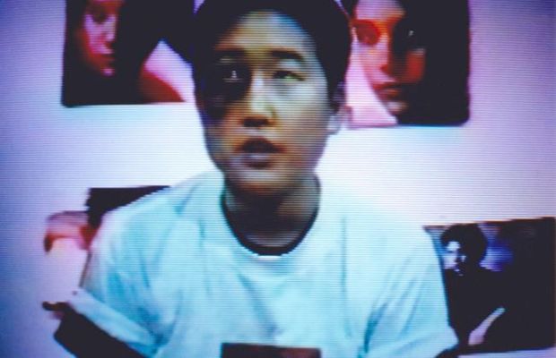 The person has light skin with a rolled up short sleeve white t-shirt with a black t-shirt underneath. They have short black hair and a big dark spot on their left eye expanding to their cheek bone. The background is white with different posters on the wall.