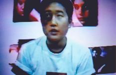 The person has light skin with a rolled up short sleeve white t-shirt with a black t-shirt underneath. They have short black hair and a big dark spot on their left eye expanding to their cheek bone. The background is white with different posters on the wall.