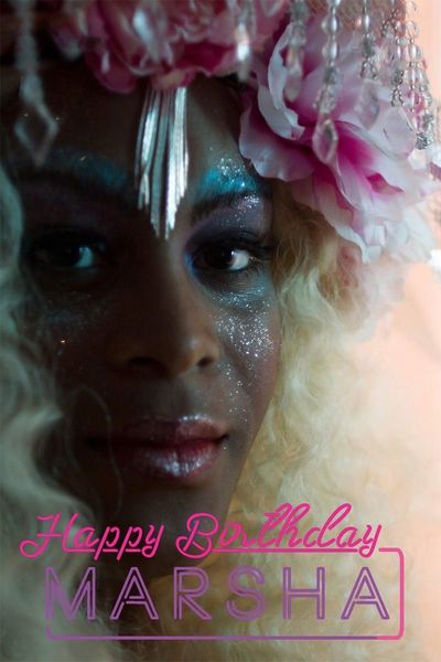 At the bottom of the image is a cursive writing of “Happy Birthday Marsha”. “Happy Birthday” is written in pink and “Marsha” is written in purple. Above that there is a face with brown skin. They have their makeup done and big blonde curly hair and a flower crown with jewels on it. They have a bunch of glitter over the face and a glittery glossy pink lip. Jewelry comes down from the crown and rests on the center of the forehead between their eyebrows. They stare into the camera with a very slight smile.