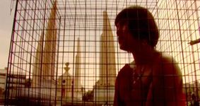 A Southeast Asian person sits in a metal cage outside of the Democracy Monument in the city center of Bangkok, Thailand. Their hair is in a short bob with a fringe and they wear an off the shoulder dress decorated with a large necklace and earrings.