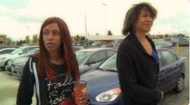 Two genderfluid people walk through a parking lot together, possibly a parent-child relationship. One appears older and is a light skinned Black person wearing a black blazer and blue shirt with a dark brown bob hairstyle, and the other is a younger dark skinned Black person wearing a black zipper hoodie and a dark graphic t-shirt, holding a coffee cup.