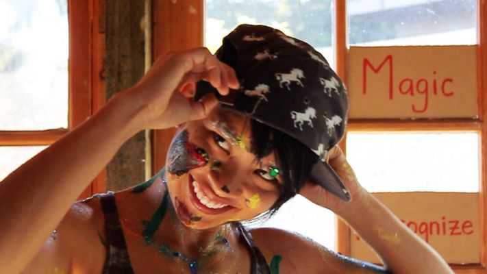 A person with tan skin is covered in paint with short hair. They are wearing a black bucket hat with white unicorns on the hat. They are posed in front of a wooden wall with windows shining in light. They stare up past the camera with a diabolical smile.