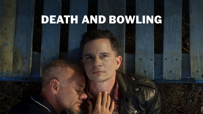 Two people are lying on a blue block palette. The one on the right stares up contemplatively past the camera as the one on the left lies on the right one's shoulder, their eyes closed, cuddling the person on the right with their hand on their chest. The person on the right has short brown hair with faded sides. They are wearing a red and black flannel and a black leather jacket. The person on the left has their eyes closed, a goatee and light brown short hair with faded sides. They are wearing a navy blue polo with a thin white line at the edge of their collar.