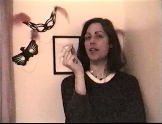 A person is mid-sentence against an off white wall. The person has light skin and shoulder length straight brown hair. They are wearing a black top with a white collar and half their right arm crossed over their body with their left hand resting on it up near their face. In the background there are black masks on the wall and a picture frame behind them.