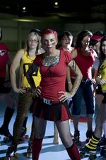 Eight people pose with one person in the forefront. The person in the front has light skin, tattoos covering their chest and arms. They have a red short sleeve t-shirt, a red and black pleated skirt, a red belt with the belt buckle that says "evil", and knee high socks. They are posed with their hands on their hips, they have red hair pulled back, a piercing under their lip, and red and black striped face paint on either cheek. The person behind them to the left is posed in the same way with light skin, blonde hair and glasses. They are wearing a yellow tank top and yellow cheetah print pants with roller skates on. There are varying people behind them posing, some with red shirts and some in yellow, most in roller skates. The background is dimly lit with the focal point of the light on the group. The floor is blue with yellow lines on the floor and some red circles.