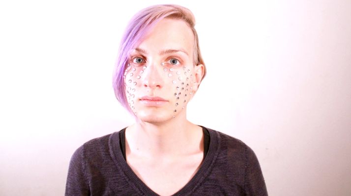 A person in front of a white background with light skin and short purple hair. They have a bunch of small diamonds on their face from their eyeline down to their jaw line. They have a black sweater on.