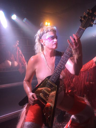 Peaches performing a guitar solo live at a concert venue. Peaches is a white genderqueer person with a blonde mullet, artistic colourful makeup on the eyebrows, cheekbones and lips, and wears a theatrical costume that matches two background dancers in the photograph.