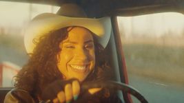 Ponyboi (River Gallo) drives a truck amd smiles with the sun shining on their face. Their left hand is on the steering wheel with blue nails and black fingerless gloves. They have long brown curly hair, light brown skin, and are wearing a beige cowboy hat, gold hoop earrings, and a black leather jacket.