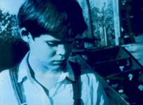 A close up of a light-skinned child gazing toward the ground. They appear from the shoulders up, have short dark hair, and are wearing a collared shirt with overalls. They are outside in front of bushes and a staircase.